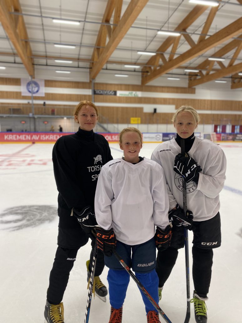 En jente i svart  og to jenter i hvite  hockeyskjorter står på isflaten med skøyter og hockeykølle