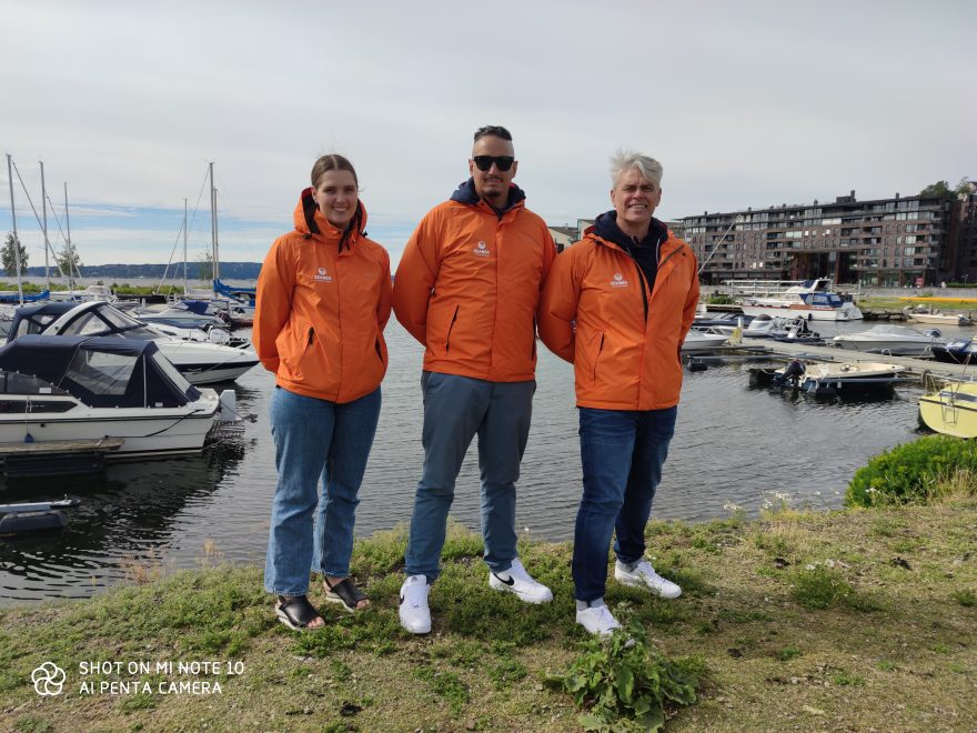 En kvinne og to menn i oransje jakker i en båthavn